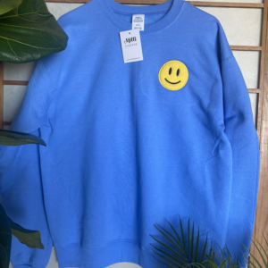 The embroidered yellow smiley face in blue sweatshirt hang on the wall with plants background and Millithreads tag in shirt.